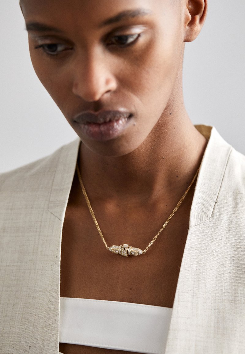 Zelta kaklarota ar teksturētu piekariņu, kas papildināts ar caurspīdīgiem akcentiem, valkāta virs gaiši bēša žaketes un balta krekla.