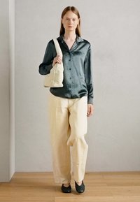 Camisa de cetim verde escuro com botões à frente, calças largas creme, sapatos pretos de slip-on e uma bolsa creme com uma textura suave.