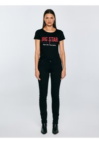 Mujer de pie, mirando al frente, con una camiseta negra de "BIG STAR" y vaqueros negros, junto con botines negros, sobre un fondo claro y liso.