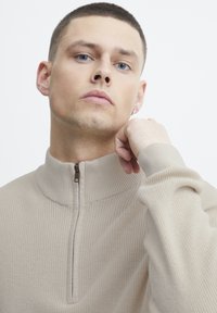 Pull en beige à fermeture éclair avec texture côtelée et col montant. Doté d'une fermeture éclair d'un quart de longueur et de poignets ajustés. Tissu lisse avec un éclat subtil.