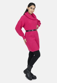 Stickad oversized tröjklänning i stark rosa med en hög krage, bältad med ett svart bälte med hjärformad spänne, tillsammans med svarta leggings och stövlar.