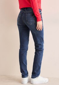 Vaqueros de mezclilla azul oscuro con un corte ajustado, con bolsillos traseros y un suave desgaste. Combinados con zapatillas blancas para un look casual.