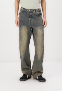 Világoskék, bő szárú farmernadrág, amely elhasználódott hatású, denim anyagból készült, és varrott részletekkel rendelkezik. Fekete sportcipővel viselve.