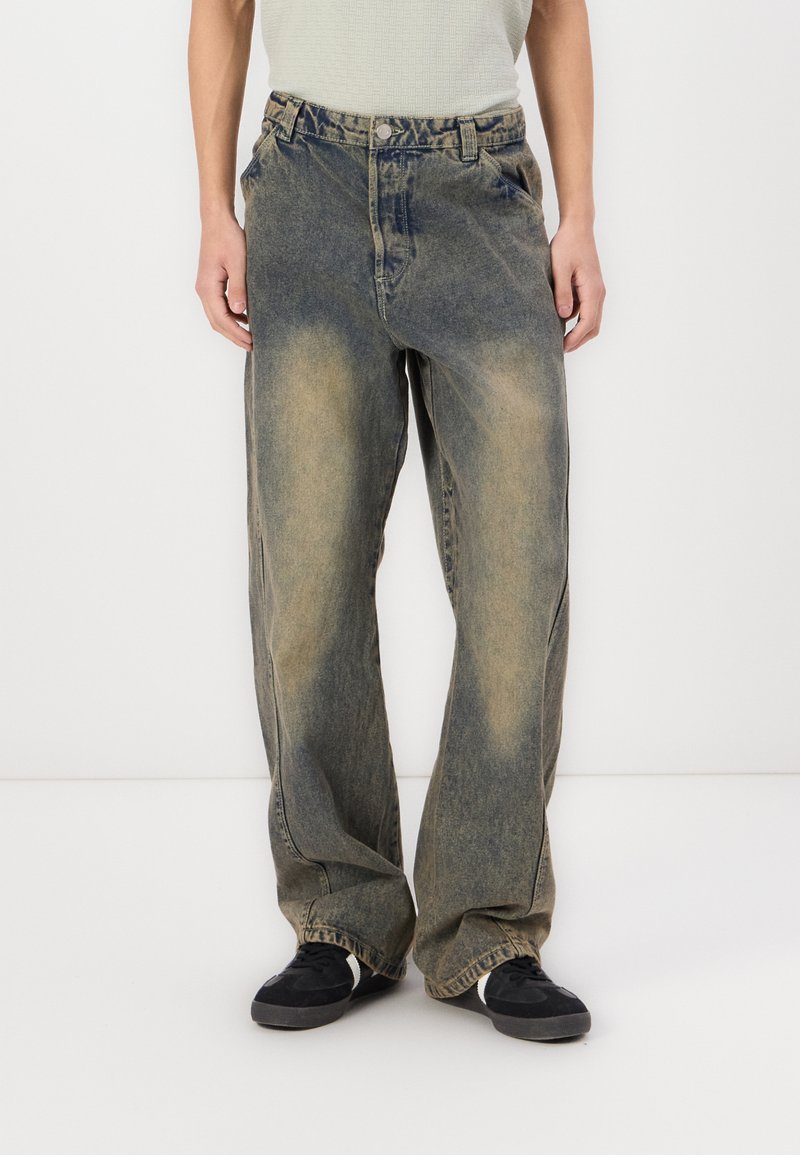 Világoskék, bő szárú farmernadrág, amely elhasználódott hatású, denim anyagból készült, és varrott részletekkel rendelkezik. Fekete sportcipővel viselve.