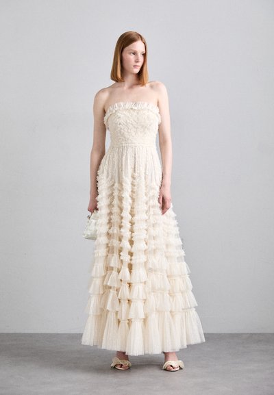 Robe crème sans manches avec un design à volants superposés, en tissu texturé et une silhouette fluide. Accessoirisée avec un petit sac à main blanc.