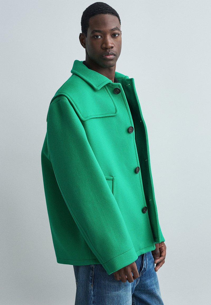 Chaqueta de mezcla de lana verde con cierre de botones, gran cuello y bolsillos laterales. Presenta un corte relajado y una textura suave. Jeans de mezclilla visibles.