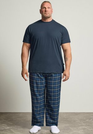Homme aux cheveux courts et tatoué, debout, portant un t-shirt bleu marine, un pantalon à carreaux bleu et des chaussettes blanches sur un fond uni.