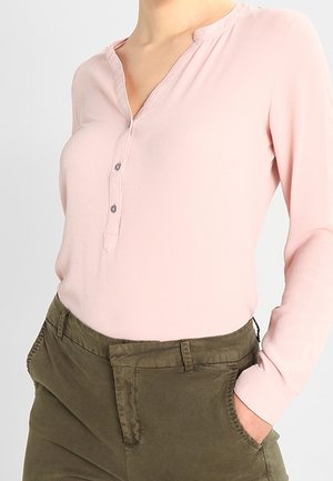 Nainen pukeutuu vaaleanpunaiseen pitkähihainen henley-paitaan, joka on tungettuna oliivinvihreisiin korkeavyötäröisiin housuihin ja pitää kättään taskussa.