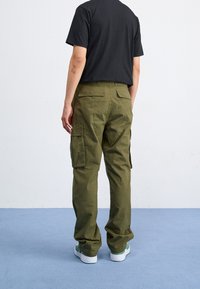 Oliwkowe spodnie typu cargo z bocznymi kieszeniami, luźnym krojem i wytrzymałego bawełnianego materiału. Noszone z czarnym T-shirtem i białymi trampkami.