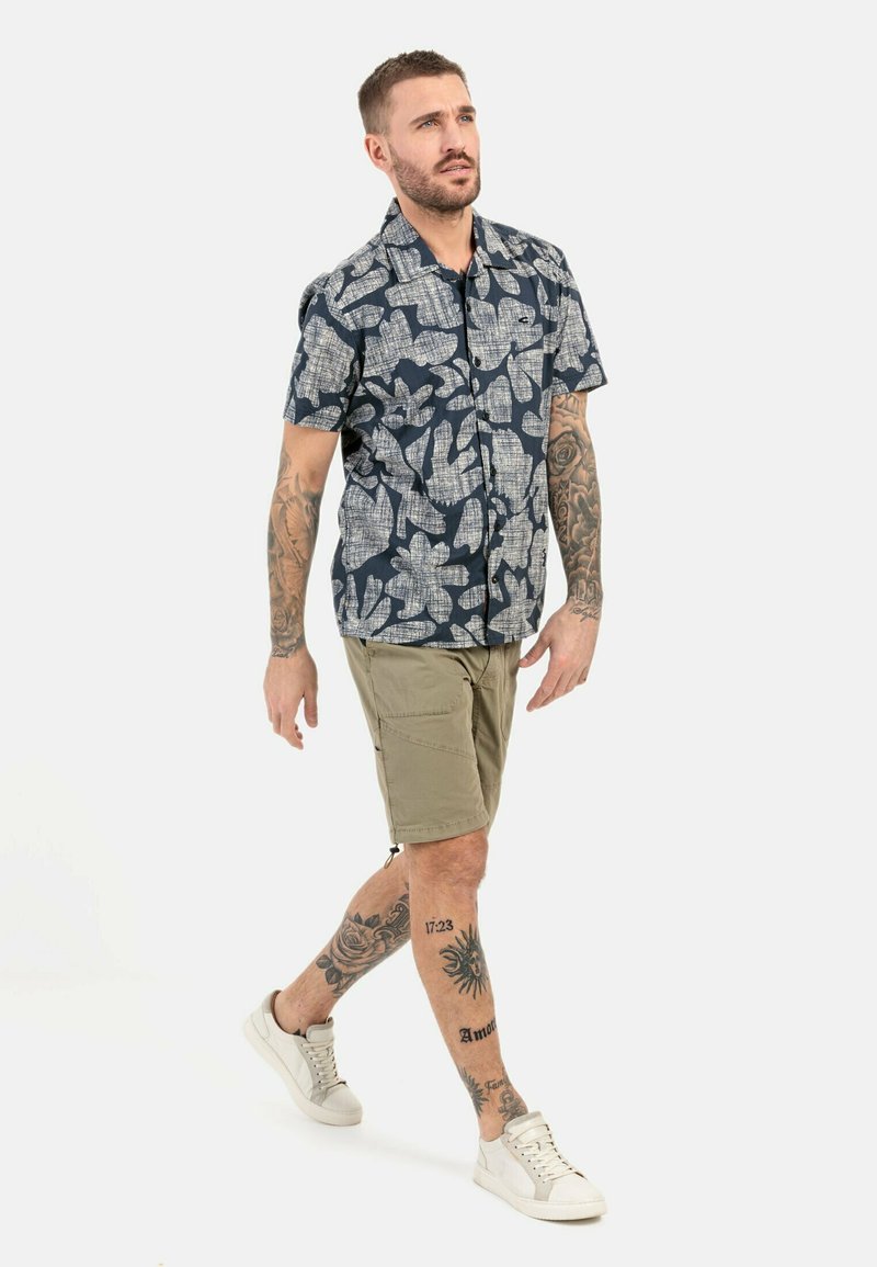 Kurzarmliges, navyblaues Hemd mit einem weißen Blumenmuster, kombiniert mit beigen Cargo-Shorts und weißen Turnschuhen. Glattes Material, lässiges Design.