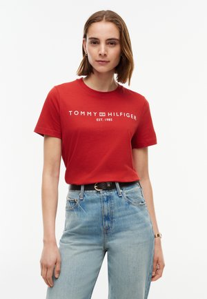 Giovane donna che indossa una t-shirt rossa Tommy Hilfiger e jeans azzurri chiari, in piedi con la mano rilassata lungo il corpo davanti a uno sfondo semplice.