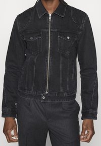 Svart denimjacka med dragkedja fram, spetsig krage, två bröstfickor och sidofickor. Har kontrasterande sömmar och en figurnära design.