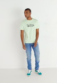 Gaiši zaļš T-krekls ar melnu "Pepe Jeans" logo, kombinēts ar izbalējušiem zilajiem džinsiem un zaļajiem sporta apaviem. Minimalistisks dizains ar ikdienišķu piegriezumu.