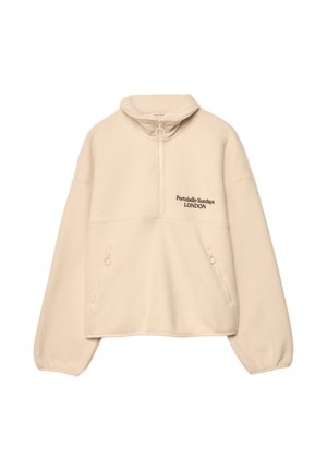 Osoba nosząca kremowy polar zapinany na zamek z napisem „Portobello Sundays London”, dżinsowe szorty oraz czarne skórzane kozaki za kolano na jednolitym tle.