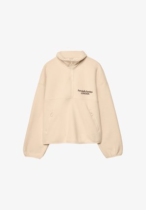 Persoon draagt een crèmekleurige fleece met rits met de tekst "Portobello Sundays London", spijkershorts en zwarte leren laarzen tot aan de knieën tegen een effen achtergrond.