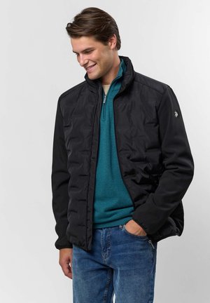 Mand smilende og kigger ned, iført sort quiltet jakke over teal kvart-zip sweater og blå jeans, hænderne i lommerne.