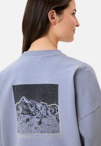 Helles blaues Sweatshirt mit einem schwarz-weißen Berggrafik auf der Rückseite, darunter der Schriftzug "TOGETHER" in goldenen Buchstaben.