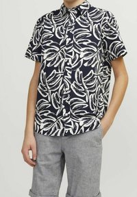 Camicetta a maniche corte con bottoni di colore blu navy e motivo astratto a foglie bianche, realizzata in tessuto leggero. Abbinata a pantaloni corti grigi chiari con risvolti.