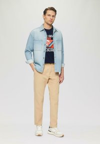 Lichtblauwe spijkerjas over een marineblauw graphic t-shirt, beige chino's en witte sneakers met zwarte accenten; een casual en veelzijdige outfit.