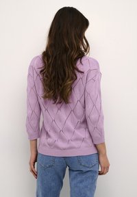 Lila stickad tröja med diamantmönster, trekvartslånga ärmar och ribbat avslut. Bärs med blå denimjeans.