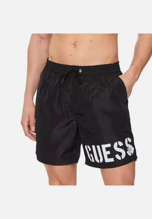 Uomo che indossa pantaloncini da nuoto neri con il logo bianco "GUESS" sulla gamba sinistra, vita elastica e coulisse, con le mani nelle tasche.