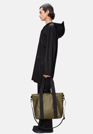 Svart vattentät jacka med huva, svarta byxor och bär en olivgrön tygpåse med svarta handtag, står upprätt.