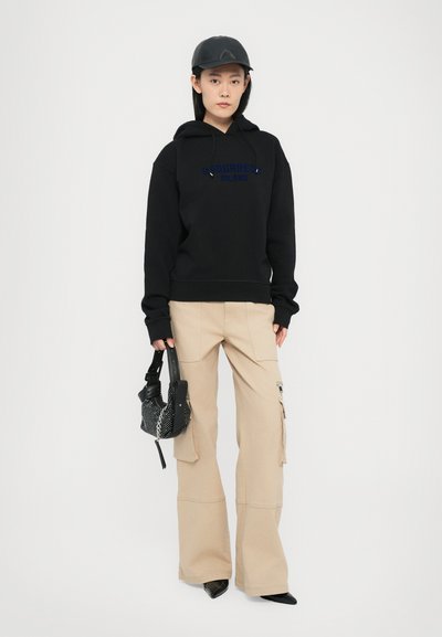 Hanorac negru cu logo albastru, pantaloni cargo bej cu buzunare mari, ținând o geantă cu un model alb și negru, purtând o șapcă neagră și pantofi ascuțiți.