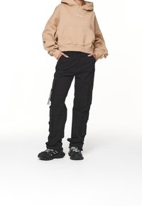 Person som bär en beige croppad hoodie och svarta cargo byxor med händerna i fickorna, stående på vit bakgrund.