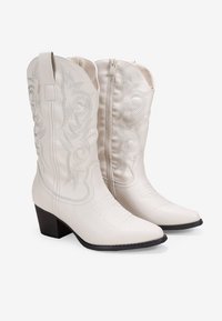Botas de cowboy de cuero blanco con costuras decorativas plateadas y punta afilada. Cuentan con un tacón cuadrado y una cremallera trasera para facilitar su uso.