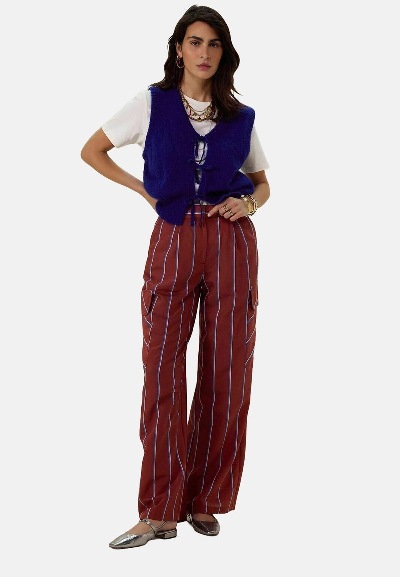 Gilet en tricot bleu sur un t-shirt blanc, associé à un pantalon large rayé marron. Des chaussures plates argentées et des colliers superposés complètent la tenue.