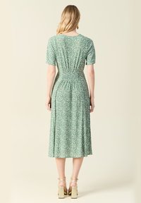 Zielona midi sukienka w kwiaty z krótkimi rękawami, marszczonym tyłem i rozkloszowanym dołem. Strukturalny materiał z białymi wzorami w kwiaty. Złote sandały na obcasie.