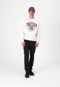 Crème kleurige crewneck sweatshirt met "TOMMY JEANS" grafische print, gecombineerd met zwarte jeans en zwarte sneakers. Een blauwe pet maakt de outfit compleet.