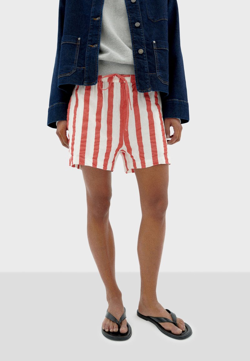 Shorts rayés rouges et blancs avec une taille élastique et un cordon de serrage, associés à un sweat-shirt gris sous une veste en denim, et des tongs noires.