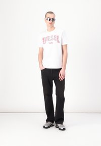 Jovem a usar óculos de sol refletivos, t-shirt branca da Diesel Core com estampa floral, jeans pretos e ténis prateados, de pé com as mãos nos bolsos.