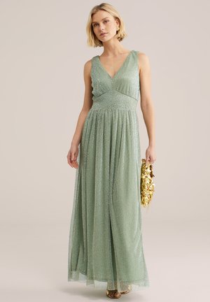 Naine, kes kannab helkiva helerohelise varrukateta pikkade kleiti, hoides kätt särava kuldse litritega käekotil, seisab ühevärvilise tausta ees.