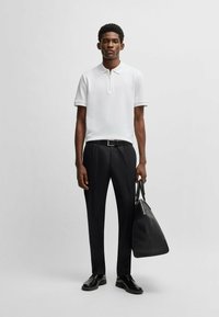 White short-sleeve polo with a zip collar, paired with black tailored trousers and black shoes, holding a large black leather bag.