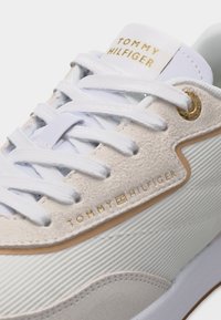 Κοντινό πλάνο λευκού αθλητικού παπουτσιού Tommy Hilfiger με χρυσό λογότυπο, λευκά κορδόνια και υφασμάτινη υφή με λεπτομέρειες στο πλάι και τη γλώσσα.