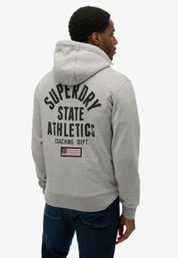 Grijze hoodie met "SUPERDRY STATE ATHLETICS" in het zwart bedrukt. Bevat een klein detail van de Amerikaanse vlag. Voorzien van geribde manchetten.