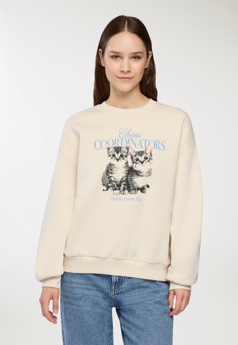 Felpa color crema con maniche lunghe, caratterizzata da una stampa grafica di due gattini e la scritta "Chaos Coordinators" in blu. Texture morbida.