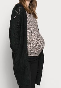 Svart stickad cardigan med öppen silhuett, prydd med diamantmönster; bärs över en rosa och gråspräcklig topp, tillsammans med svarta byxor.