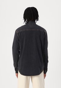 Mörk denimskjorta med långa ärmar, knapplås vid manschetterna och kontrasterande sömmar. Har en rundad fåll och en detaljerad ryggok.