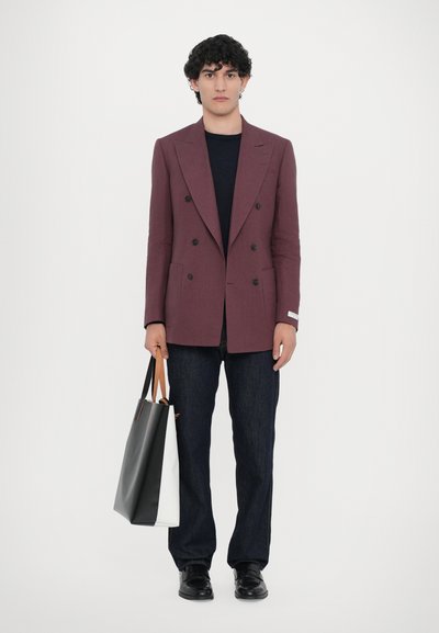 Doppelt geknöpfter bordeauxfarbener Blazer, kombiniert mit einem marineblauen Hemd, dunklen Jeans und schwarzen Schuhen. Tragend eine schwarz-weiße Tragetasche.