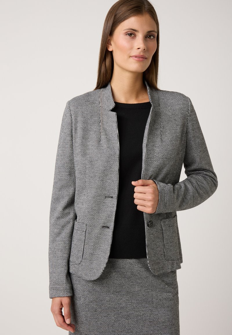 Grijze houndstooth blazer met een rechte snit, sluiting met één knoop en twee voorzakken, gedragen over een zwart shirt. Glad textuur.