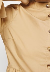 Blusa beige con bottoni e colletto alto, con una vita arricciata e dettagli di cucitura sottili, realizzata in tessuto leggero.