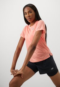 Γυναίκα σε κοραλί κοντομάνικο πουκάμισο και μαύρες βερμούδες που τεντώνει τα πόδια της με τα χέρια δεμένα πάνω στο γόνατο, μπροστά σε λευκό φόντο.