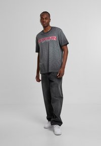 Šedé tričko s krátkým rukávem a výrazným červeným logem "True Religion", v kombinaci s volnými tmavě šedými džíny a bílými teniskami.