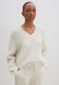 V-Ausschnitt-Pullover aus weichem, flauschigem weißen Material; mit überschnittenen Schultern und einem lockeren Schnitt, kombiniert mit hellbeigen Jogginghosen mit Kordelzug.