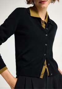 Sort cardigan med V-hals og knaplukning. Har en mørkegrøn krave og kontrasterende manchetter. Blød tekstur, tætsiddende design.