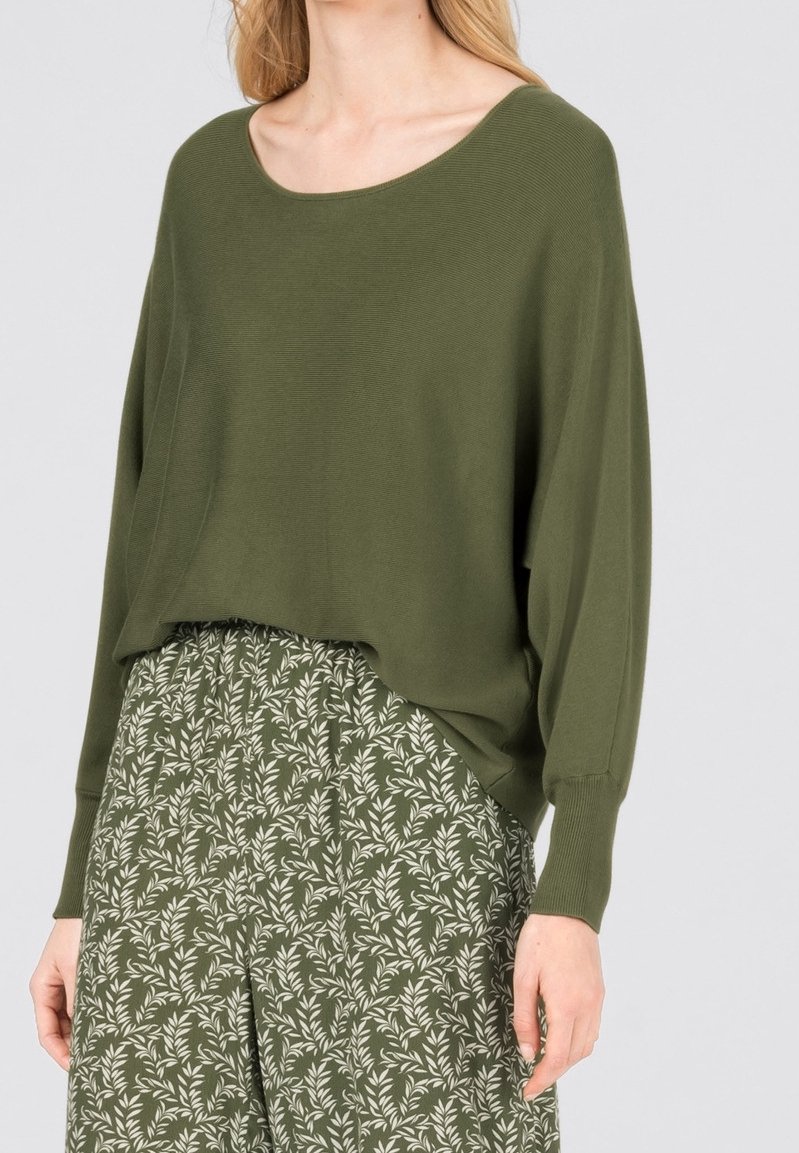 Femme portant un pull ample vert olive à manches longues et une jupe imprimée vert olive et blanche.