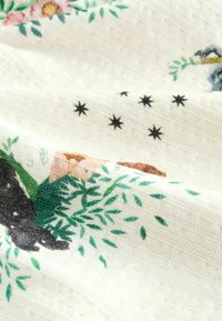 Gemusterter Stoff mit floralen und sternförmigen Motiven in Grün, Schwarz und Pfirsich auf einem cremefarbenen Hintergrund, der eine strukturierte Webart zeigt.